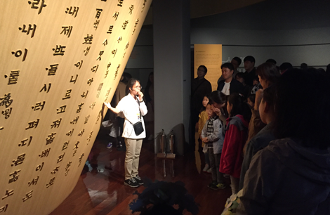 어린이 해설사가 한글박물관 전시관의 전시물을 가리키며 마이크를 손에 쥔 채 수십명의 관람객에게 해설하고 있다.