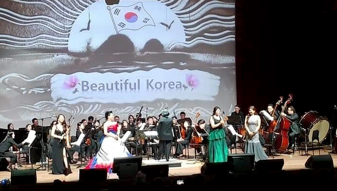 무대 뒤편으로 ‘Beautiful Korea’라 적힌 스크린이 떠 있고, 무대에는 수십명의 교향악 연주자가 지휘자의 지휘에 맞춰 연주하고 있다. 그 앞에 네 명의 여성 성악가가 드레스를 곱게 차려입고 노래하고 있다.