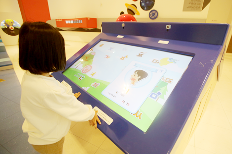 한 여자 아이가 터치스크린 앞에서 스크린에 떠오르는 한글 글자들을 바라보고 있다.