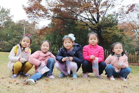 다섯명의 여자 아이들이 잔디밭 위에 쪼그려 앉아 카메라를 보며 포즈를 취하고 있다.