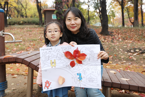 엄마와 어린이가 손에 A3정도 크기의 종이판을 들고 카메라를 응시하고 있다. 종이판 위로는 아이와 함께 찾은 낙엽들이 예쁘게 붙어있다.