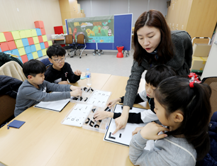 프로그램 강사가 손가락으로 판을 가리키며 두 아이에게 조언해주고 있다. 사진 좌측의 두 어린이도 무언가에 열중해 만드는 중이다.