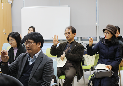 여섯 명의 수강생이 의자에 앉아 오른손을 들어 손가락을 구부리고 펼치는 수어 동작을 따라하고 있다.