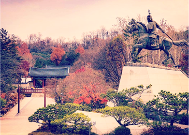 단풍이 든 낙산대 공원 풍경. 녹색의 침엽수와 단풍이 들어 주황빛이 도는 활엽수가 섞여 있다. 그 사이에 전통 한옥 건물이 보인다. 건물은 기와지붕으로 되어있다. 오른쪽에는 kf을 타고 있는 장수의 동상이 있다.