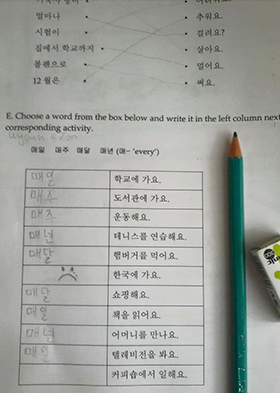 외국인용 한글 공부 책의 내용이다. 상단에는 호응이 맞는 동사를 찾아 줄로 연결하는 문제가 출제되어 있으며 하단에는 호응이 맞는 단어를 빈칸에 채우는 문제가 출제됐다. 서툰 글씨로 ‘매일’, ‘매주’, ‘매년’ 등의 단어가 칸에 적혀있다. 문제 옆에는 초록색 연필이 놓여있다.