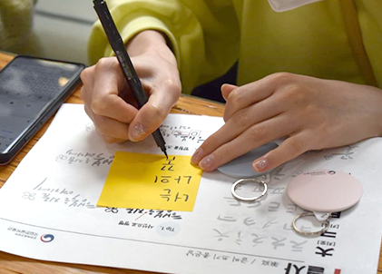 국립한글박물관에서 제공하는 한글 캘리크래피 체험을 하는 움만. 다양한 캘리그래피가 적힌 책자 위에 노란색 포스트잇을 붙이고 글씨를 연습하고 있다. 그 옆에는 한글박물관에서 제공한 사은품도 보인다. 사은품은 동그랗고 손바닥 크기만하며 어딘가에 걸 수 있게 링이 달려있다.