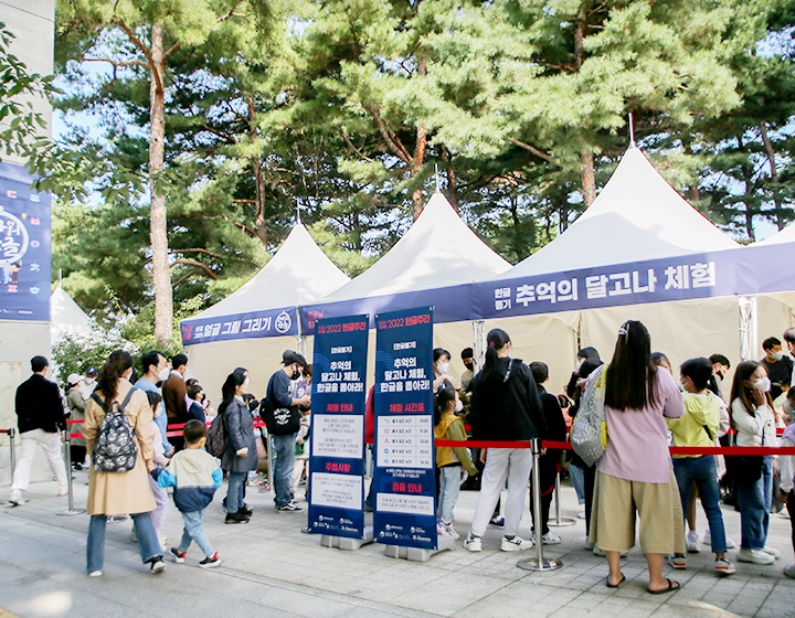 기획기사 기사 사진. 참여 행사 부스가 나란히 설치되어 있다. 그 앞에 수많은 사람이 줄을 서서 기다리고 있다. 사람들 주변에는 ‘추억의 달고나 체험, 한글을 뽑아라!’라는 문구와 함께 체험 시간표, 경품 안내 등을 적어놓은 입간판이 놓여있다. 멀리 기둥에는 ‘고마워 한글’이라고 적힌 포스터가 붙어있다.