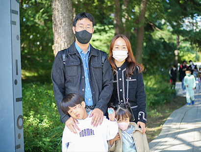 검은색 재킷을 입은 남성(아버지)과 검은색 재킷을 입은 여성(어머니)가 나란히 서 있다. 남성은 검은색 마스크를 착용하고 가방을 메고 있다. 그들 앞에는 각각 남자아이와 여자아이가 나란히 함께 서 있다.