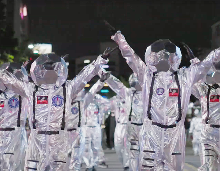 문화행사 기사 사진. 은색 우주복을 입고 머리엔 은색 축구공 모양의 헬멧을 쓴 사람들이 나란히 서 있다. 그들은 검은색 장갑을 착용하고 있으며 양손을 위로 높게 뻗고 있다.