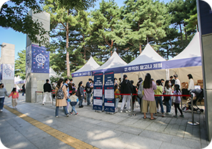 참여 행사 부스가 나란히 설치되어 있다. 그 앞에 수많은 사람이 줄을 서서 기다리고 있다. 사람들 주변에는 ‘추억의 달고나 체험, 한글을 뽑아라!’라는 문구와 함께 체험 시간표, 경품 안내 등을 적어놓은 입간판이 놓여있다. 멀리 기둥에는 ‘고마워 한글’이라고 적힌 포스터가 붙어있다.