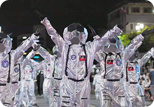 은색 우주복을 입고 머리엔 은색 축구공 모양의 헬멧을 쓴 사람들이 나란히 서 있다. 그들은 검은색 장갑을 착용하고 있으며 양손을 위로 높게 뻗고 있다.