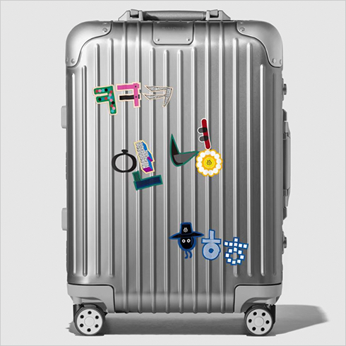 여행 가방용 한글 스티커가 붙은 은색 여행 가방 사진이다. ‘ㅋㅋㅋ’, ‘안녕’, ‘ㅎㅎㅎ’라 쓰인 스티커가 가방에 붙어있다.
