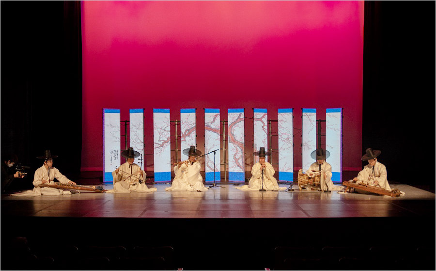 
                                        전통음악집단 샛의 공연 사진이다. 여섯 명의 단원이 무대 위에 일렬로 나란히 앉아 제각각 전통 악기를 연주하고 있다. 단원들 뒤엔 병풍 10개가 놓여있고, 그 뒤엔 빨간색 배경이 있다.