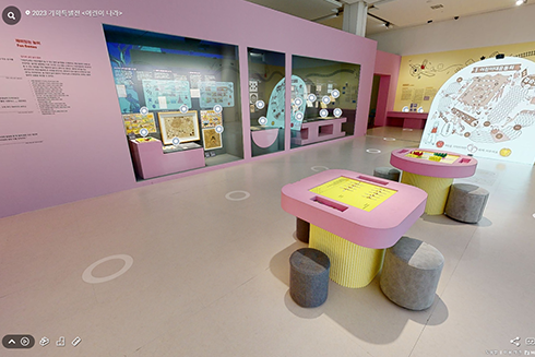 2023 기획특별전 <어린이 나라> 온라인 전시(VR) 화면이다. 분홍색과 노란색으로 만들어진 테이블 2개와 회색 의자들이 오른쪽에 놓여있고, 왼쪽엔 분홍색 벽 안에 전시품들이 놓여있다. 