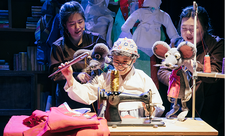 출연단체 극단 봄의 공연 사진이다. 할머니 분장을 한 여성이 재봉틀로 한복을 만들고 있다. 그 뒤엔 두 여성이 각각 쥐 형상의 인형을 들고 공연을 하고 있다.