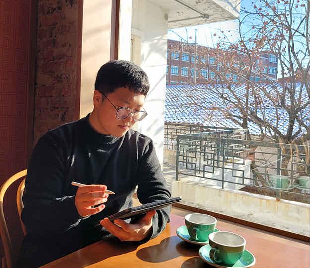 검은색 상의와 안경을 쓴 진관우 작가가 탁상에 앉아 태블릿 PC로 그림을 그리고 있다. 탁상 위엔 초록색 찻잔 두 개가 놓여있고, 탁 트인 창밖에는 나무와 건물들이 보인다.