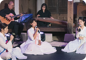 정가앙상블 소울지기의 공연 사진이다. 조선시대 가정집을 재연한 무대에서 정가앙상블 소울지기가 공연하고 있다. 앞에는 흰색 옛 의상을 입은 여성 3명이 노래하고 있고, 뒤에는 검은색 의상을 입은 남성과 여성이 각각 악기를 연주하고 있다.
