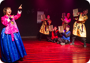 극단 가로수포엠의 공연 사진이다. 왼쪽엔 한복을 입은 여성이 양손을 들고 공연하고 있으며, 오른쪽엔 전통 복장을 한 단원들이 각자 책 한권을 들고 공연하고 있다.