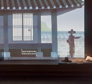 한글 노래 『한양가』를 중심 주제로 한 기획특별전 <서울 구경 가자스라, 한양가> 전시 현장 사진이다. 화면에 옛 기와집과 사람 한 명이 나와 있으며, 화면 앞엔 갓을 비롯해 옛 물품들이 전시돼 있다.