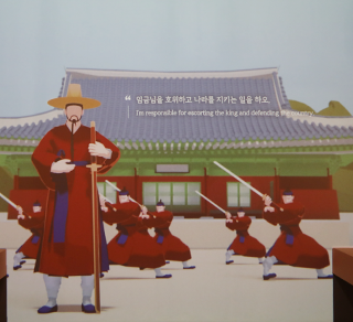한글 노래 『한양가』를 중심 주제로 한 기획특별전 <서울 구경 가자스라, 한양가> 전시 현장 사진이다. 옛 건물 앞에 빨간 옷을 입은 병사들이 검을 들고 훈련하고 있다.