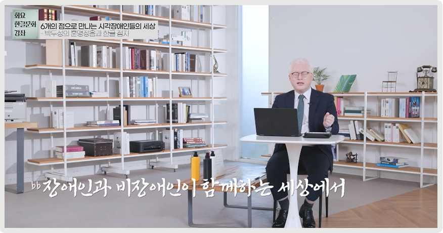 2023년 국립한글박물관 <화요 한글문화 강좌> 제9회차 강연, 김동복 한국점자도서관장의 ‘6개의 점으로 만나는 시각장애인들의 세상’ 영상 장면이다. 김동복 관장이 의자에 앉아 눈을 감은 채로 이야기하고 있고, 주위에는 책장에 책들이 꽂혀 있다.