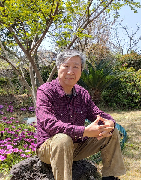 보라색 체크 셔츠를 입은 김석희 번역가가 양손을 깍지를 낀 채 바위에 비스듬히 앉아 정면을 바라보고 있다. 그의 뒤엔 꽃들과 나무, 풀이 무성히 피어 있다.