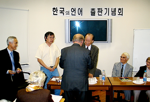 한국의 언어 출판기념회 글씨가 중앙에 있고 페이블에는 책과 물병이 있다. 1명은 앉아 있고 4명은 서 있는데 뒷모습을 하고 있는 사람이 이익섭 교수 앞에 서 있다.